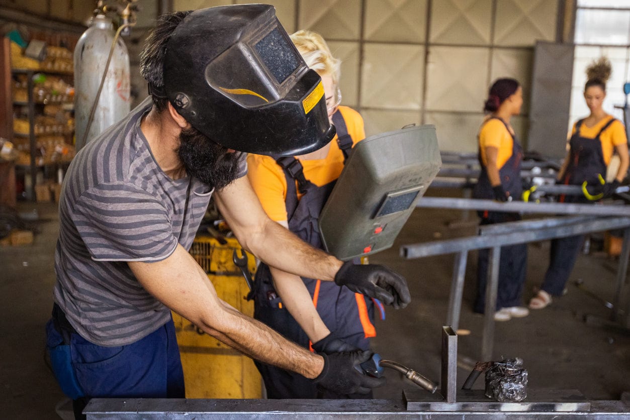 grupa spawaczy w kombinezonach słucha instruktora przy stanowisku spawalniczym